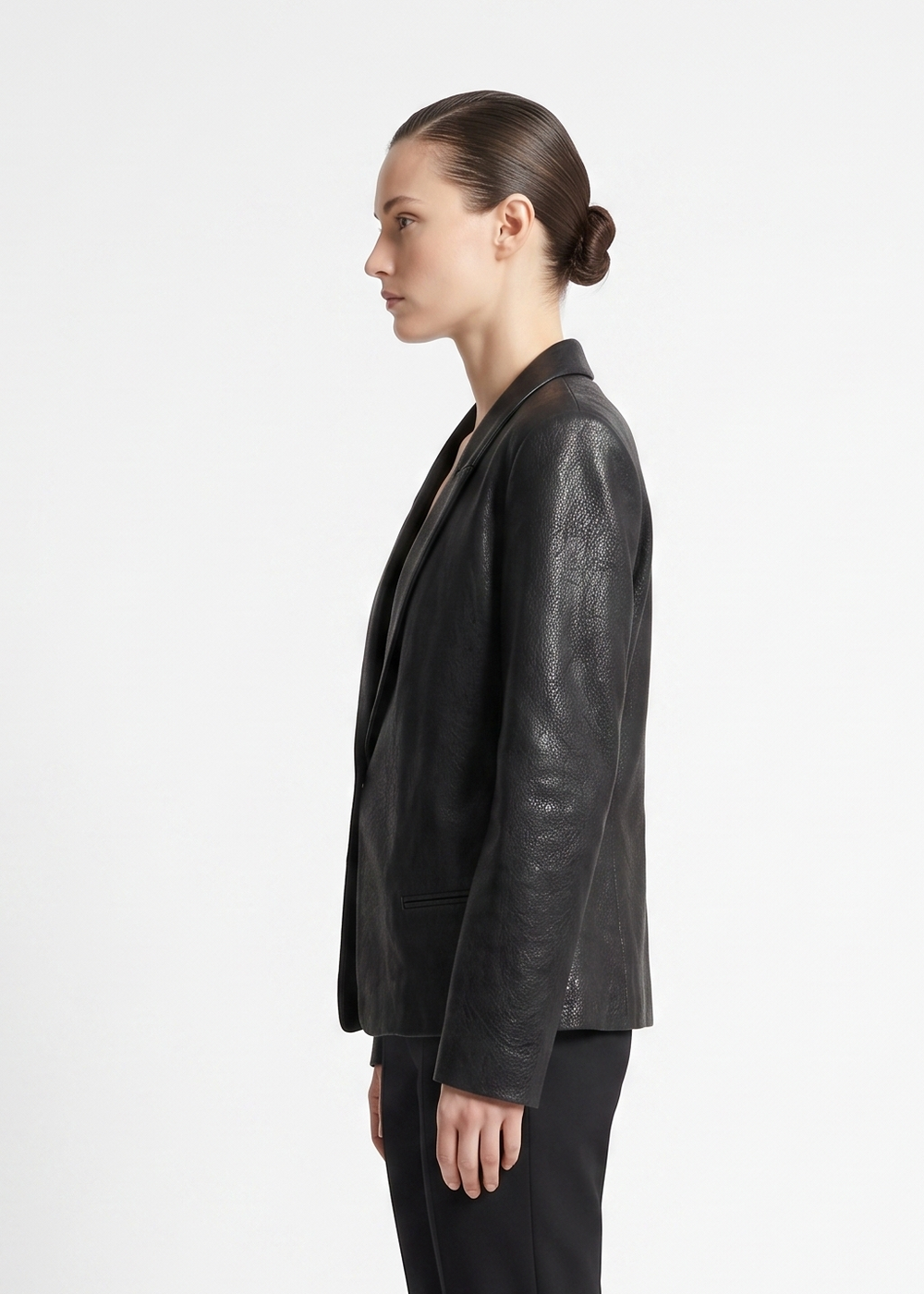 A woman shown in profile wearing a black leather blazer with a subtle tonal pattern on the sleeves, paired with black trousers, against a plain white studio background.