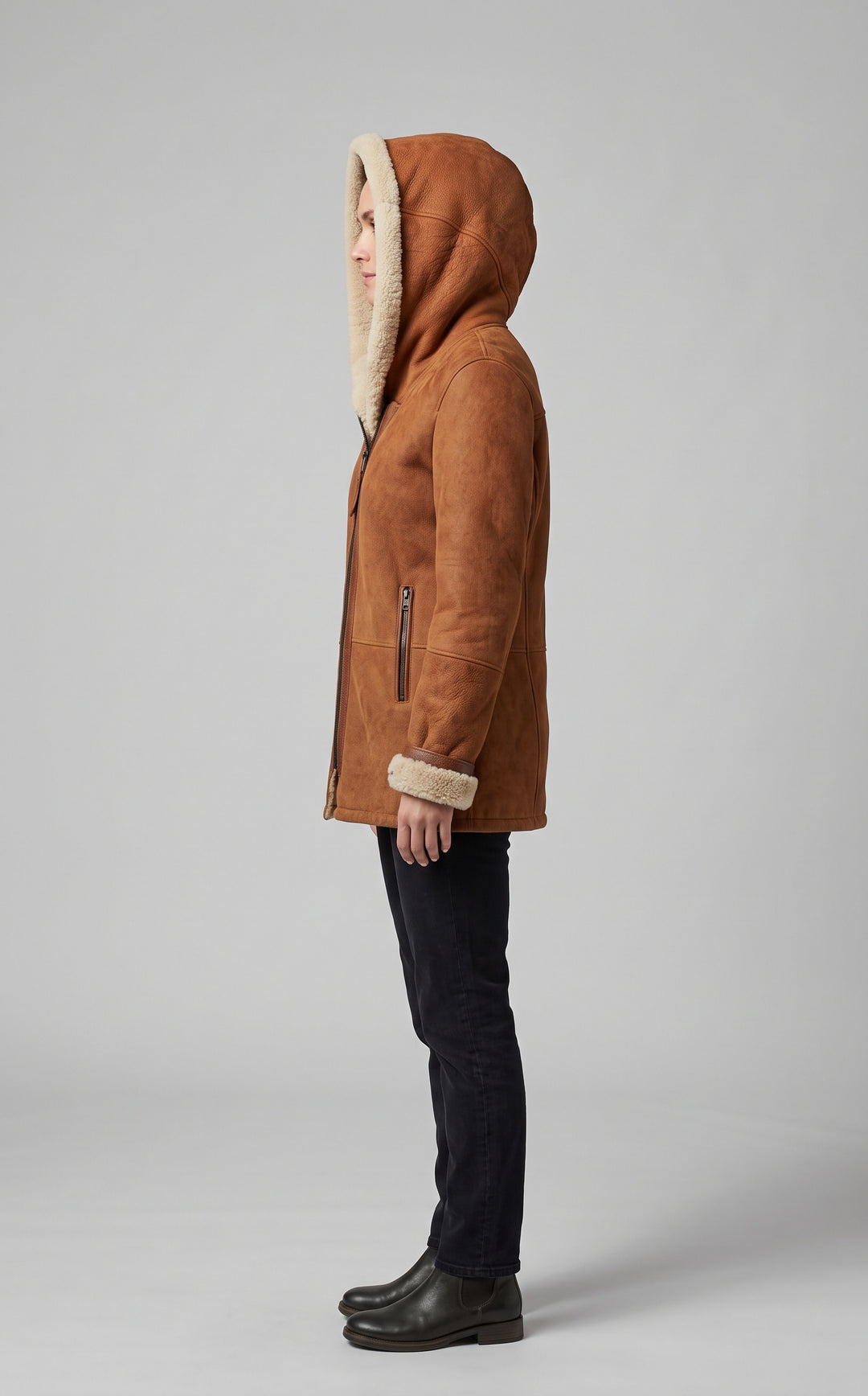 A side-profile view of a person wearing a brown suede hooded jacket with a cream-colored shearling lining, paired with dark jeans and black boots, against a neutral gray studio background.
