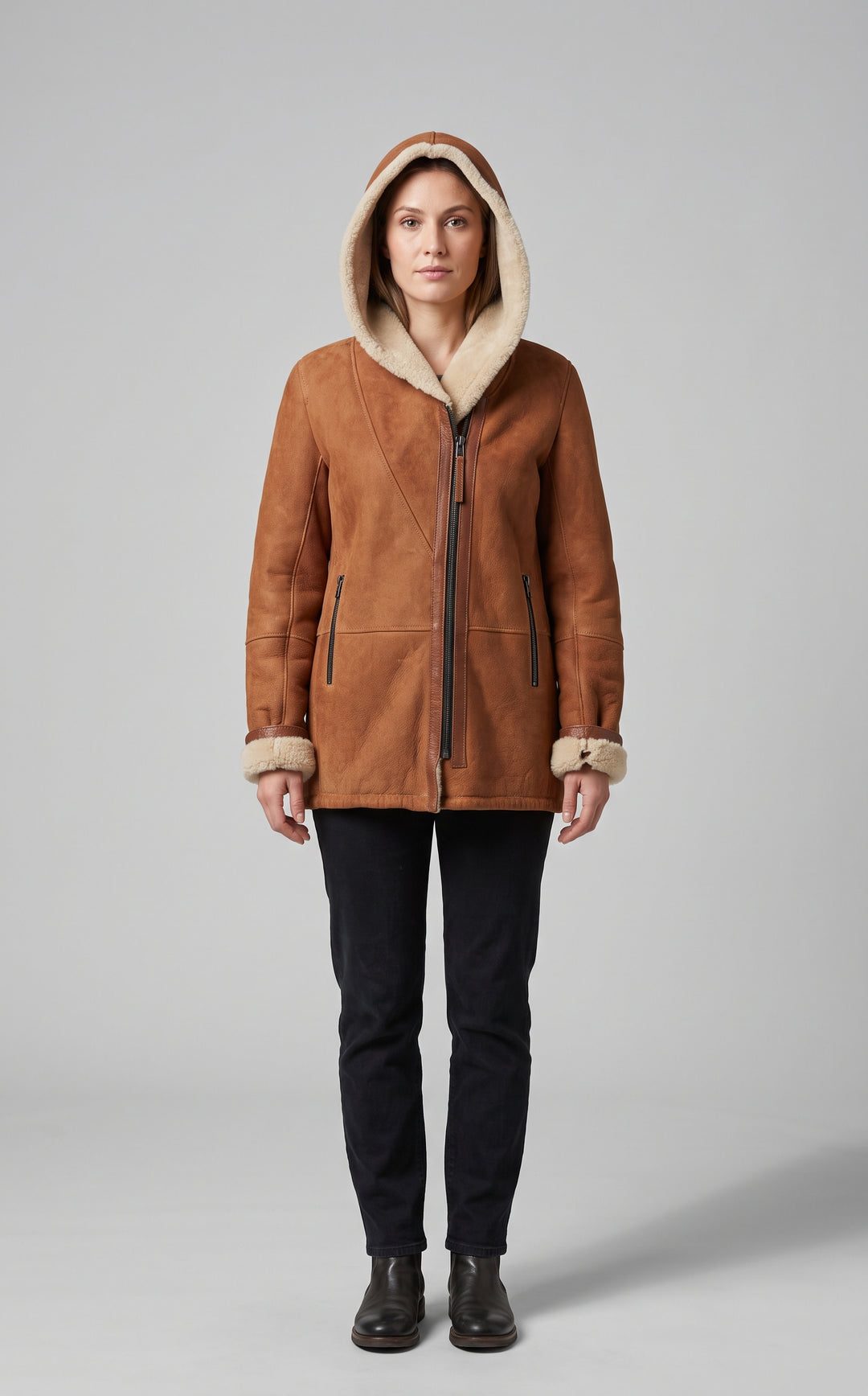 A woman stands facing forward wearing a brown suede-like hooded jacket with a cream shearling-style lining, dark pants, and dark boots against a light gray studio background.