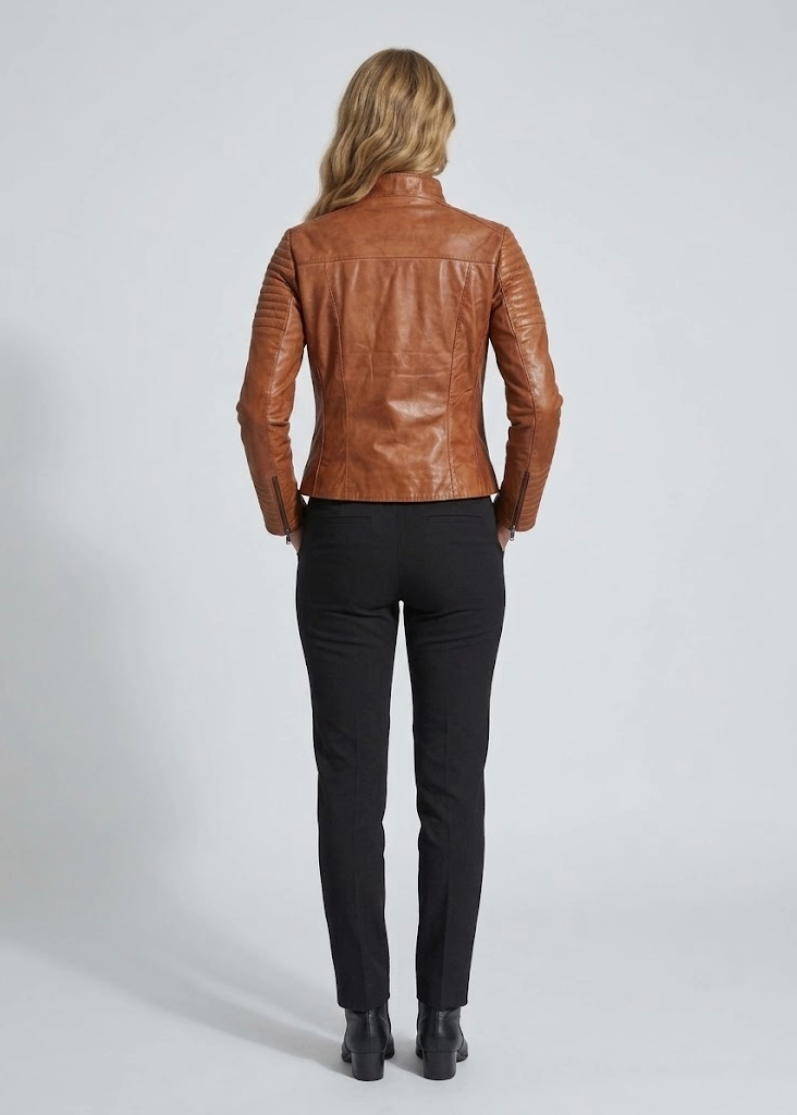 A woman viewed from behind wearing a fitted brown leather jacket, black trousers, and black ankle boots against a light gray studio background.