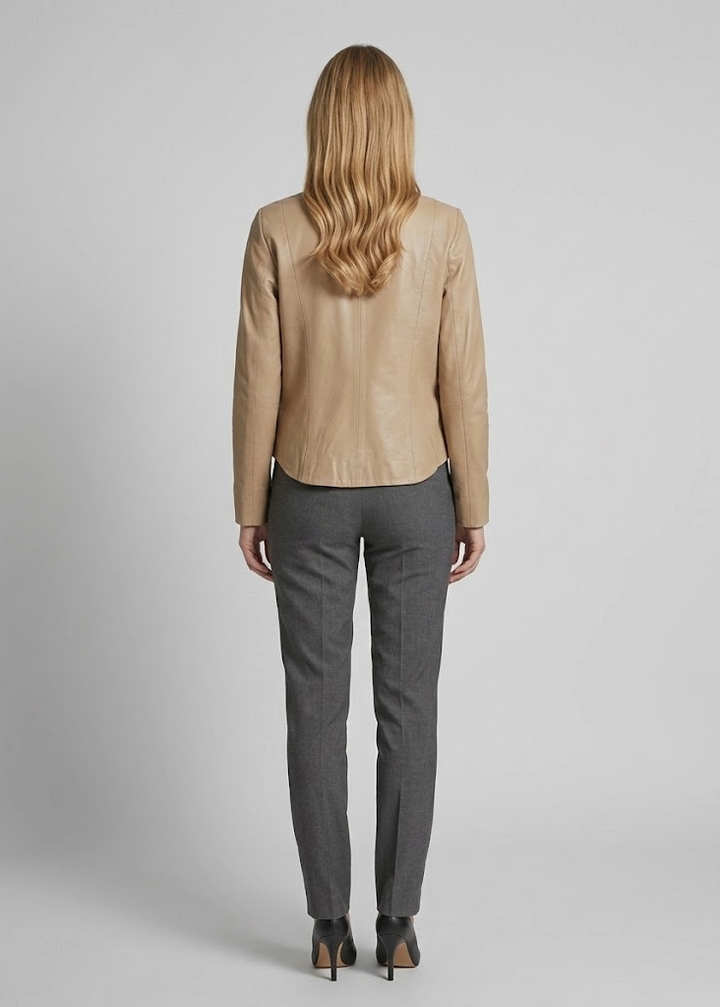 A woman viewed from the back wearing a light tan leather jacket, gray trousers, and black heels against a light gray studio background.