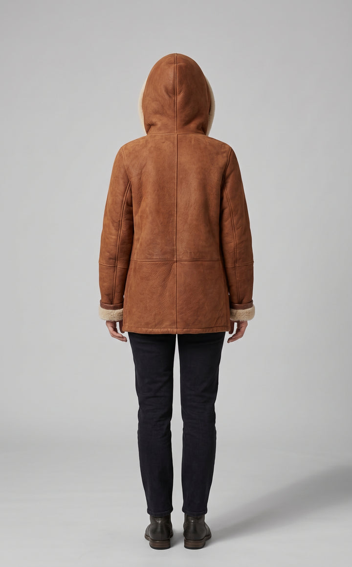 A person shown from the back wearing a brown suede-like hooded jacket with light beige trim, paired with dark pants and boots, against a plain light gray studio background.